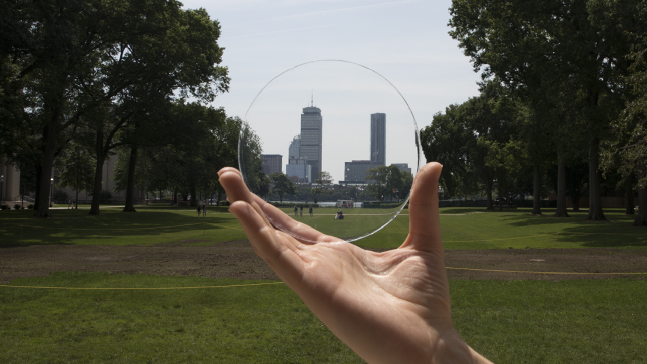 A hand holds a circular sample of Aeroshield's transparent aerogel in front of the Boston skyline.